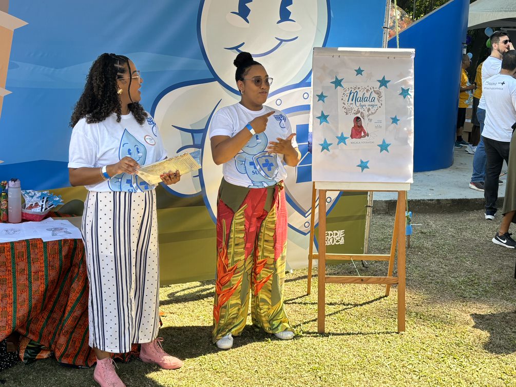 Na imagem, duas mulheres negras estão em pé. Ambas seguram livros nas mãos. Ao fundo, há um quadro com o nome 'Malala' escrito em letras grandes. As mulheres estão vestidas de forma casual, camisa branca e calça jeans. 