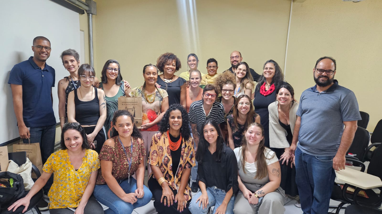 Foto com 15 pessoas em sua diversidade de genero, raça e cor, numa sala amarela. Todas estão sorrindo. 