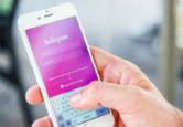 Mão masculina branca segurando um smartphone, também branco, com tela visível. A tela do smartphone em fundo rosa, tem letras brancas sobrepostas com as informações de login do Instagram