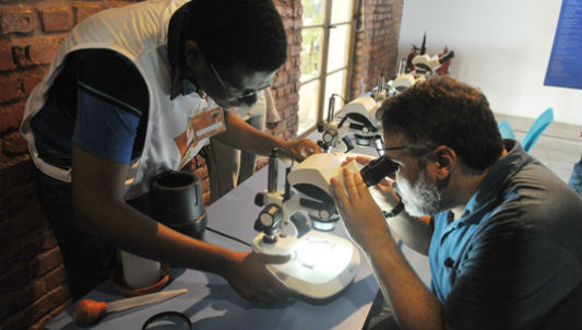 Pessoa observa algo no microscópio com ajuda de instrutor da exposição