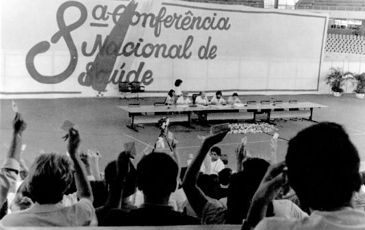 Foto antiga da 8ª Conferência: delegados de costas com mãos elevadas e mesa principal ao fundo