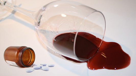 Cálice de vinho e frasco de remédio deitados sobre uma superfície branca, derramando vinho e pílulas
