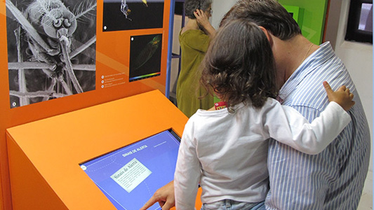 Pai com filha no colo mexe em tela interativa da exposição