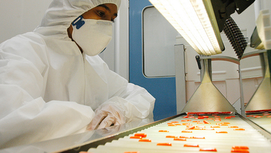 Técnico com equipamento de proteção individual observa cápsulas de remédios cor laranja em esteira de produção