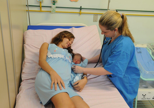 Mulher com bebê recém-nascido ao lado em uma maca recebe atenção outra mulher profissional de saúde