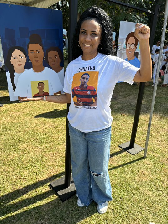 Uma mulher negra, com o cabelo preto preso e as mãos fechadas, posa em frente à sua ilustração. Na imagem, ela está acompanhada de outras duas mulheres negras. Ela veste uma camisa branca com a imagem de seu filho, um jovem negro de 16 anos que já faleceu.