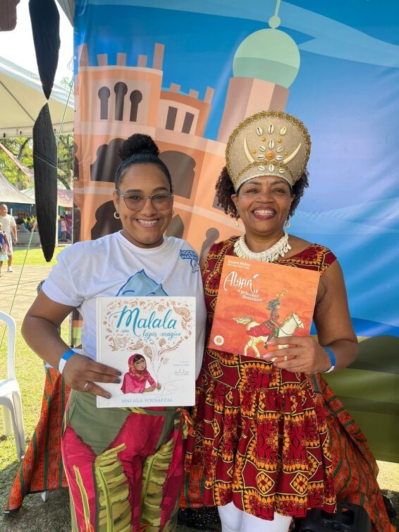 Na imagem, duas mulheres negras estão em pé. Ambas seguram livros nas mãos.