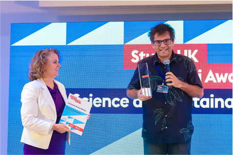 Pesquisador segura prêmio e fala em microfone. Ao lado uma mulher segura um panfleto. Ao fundo tem uma imagem azul com o escrito: Study UK Alumni Awards