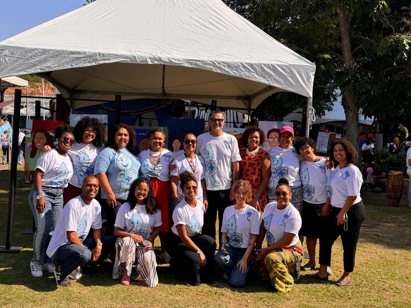 Um grupo de pessoas está posando em fila, todas vestidas com camisas brancas estampadas com "Fiocruz pra Você 2024". Ao fundo, uma tenda branca exibe placas ilustrativas que mostram rostos diversos, com variações de tons de pele, tipos de cabelo e estilos de roupa.