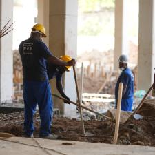 Quatro homens de uniforme azul e capacete amarelo cavam buracos em meio a muitas colunas de um prédio em construção.