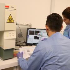 Um pesquisador e uma pesquisadora, vestindo jalecos azuis, examinam resultados de uma amostra em um computador, por meio de um equipamento de análise biomolecular.