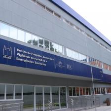 Foto da fachada do Bloco 2 do Centro de Pesquisa. É um prédio baixo, de cor cinza, com muitas janelas e portas de vidro. Sobre uma faixa em azul, estão os logos da Fiocruz, do IOC e da Ensp, ladeando o nome do centro.