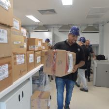 Um homem de capacete e blusa azuis carrega uma caixa de papelão em um laboratório do Centro de Pesquisa. À sua esquerda, há uma pilha de cerca de dez caixas sobre uma bancada.