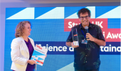 Foto de pesquisador recebendo o prêmio de uma mulher e segurando um microfone para falar em fundo azul