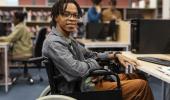 Estudante negro de dreads no cabelo, sentado em uma cadeira de rodas. Ao fundo uma biblioteca