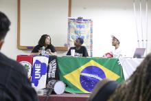 Três jovens falam ao público detra´s de uma mesa com bandeiras do Brasil, de MG, da UNE, do MST e do Levante Popular da Juventude.