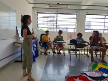 Jovens sentados em carteiras em sala de aula assistem exposição de uma jovem em uma sala de aula. Há bandeiras de movimentos sociais, do Brasil, e um estandarte do ViverSUS.