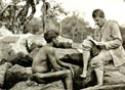 Foto antiga, em preto e branco, mostra pesquisador fazendo anotações diante de um índio