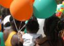Grupo de pessoas, parte de uma multidão, caminha de costas. Uma menina segura balões coloridos
