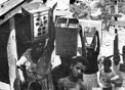Foto antiga, em preto e branco, mostra mulheres carregando latões de água na cabeça e crianças olhando para a câmera