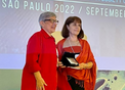 Aldina Barral vestindo blazer longo vermelho, recebendo medalha. ao fundo apresentação do evento em roxo com dizeres brancos.
