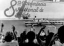 Foto em preto e branco mostra palco da Oitava Conferência, com mãos levantadas, em voto, vistas de costas