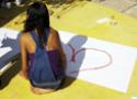 Menina faz o desenho de um coração num papel apoiado sobre o chão
