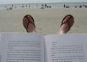 Livro aberto em primeiro plano, com pés em sandálias e paisagem de praia ao fundo