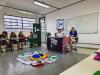 Jovens sentados em carteiras em sala de aula assistem exposição de duas jovens atrás de uma mesa. Há bandeiras de movimentos sociais, do Brasil, e um estandarte do ViverSUS